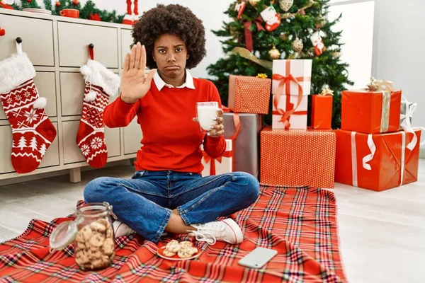 若いですアフリカ系アメリカ人女性とともにアフロ髪食べるペストリーによって座ってクリスマスツリーでオープン手を行います停止標識で真剣で自信を持って式 防衛ジェスチャー — ストック写真