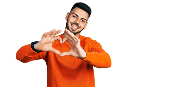 ひげを生やした若いヒスパニック系の男性は 手でハートシンボルの形をして愛に笑みを浮かべてカジュアルなオレンジのセーターを着ています ロマンチックなコンセプト — ストック写真