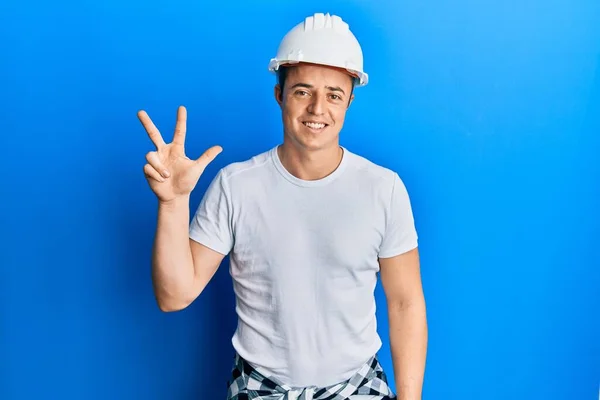 英俊的年轻人身穿建筑工人制服 头戴安全帽 三指一指 面带微笑 自信而快乐 — 图库照片