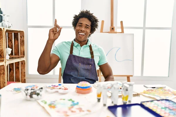若いアフリカ系アメリカ人の男性がアートスタジオのテーブルの上に座り 成功したアイデアで指を指しています 興奮して幸せよ 第1位 — ストック写真
