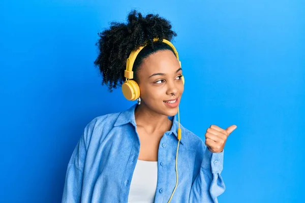 年轻的非洲裔美国女孩用耳机听着音乐 面带微笑 面带微笑 并竖起大拇指指向一边 — 图库照片