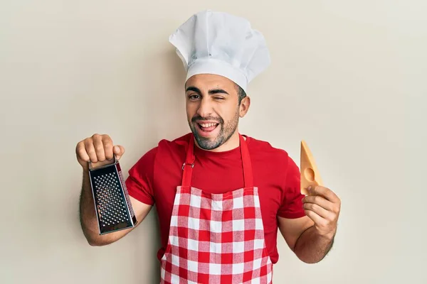 Junger Hispanischer Mann Kochuniform Reibe Und Käse Zwinkernd Die Kamera — Stockfoto