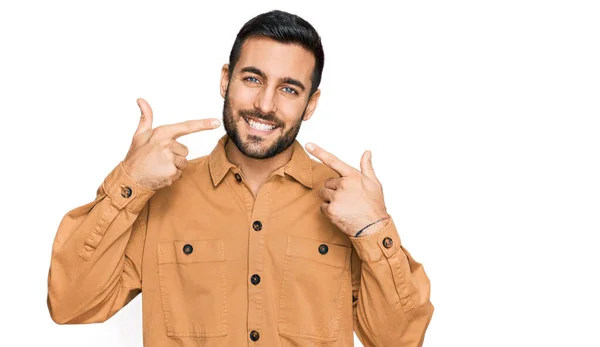 若いヒスパニック系の男は陽気な笑みを浮かべてカジュアルな服を着て指の歯と口を指しています 歯の健康の概念 — ストック写真