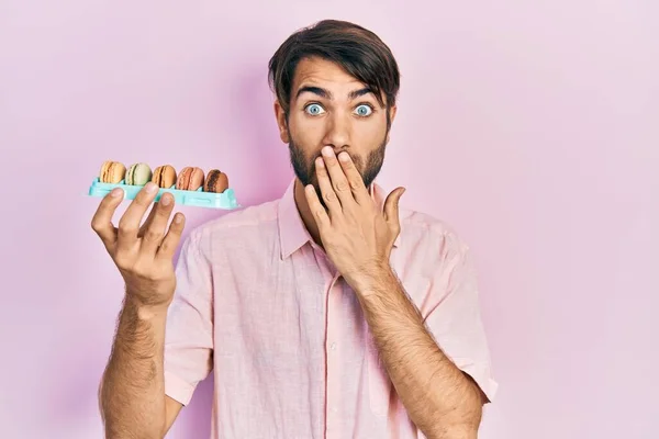Young Hispanic Man Holding Delicious Macrons Pastries Covering Mouth Hand — Stock Photo, Image