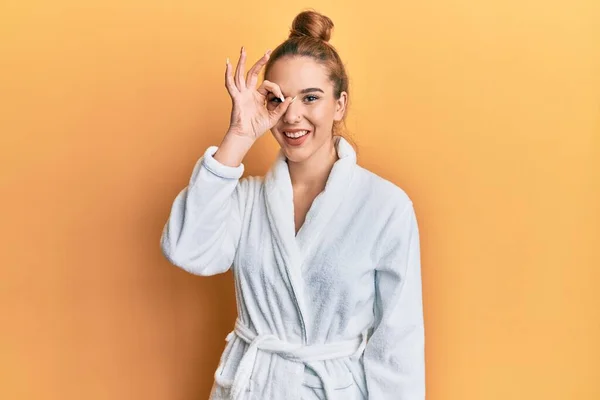 Young blonde woman wearing bathrobe doing ok gesture with hand smiling, eye looking through fingers with happy face.