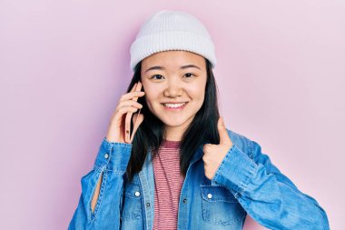 Genç Çinli kız akıllı telefonla konuşuyor mutlu ve olumlu gülümsüyor, başparmağıyla mükemmel ve onay işareti yapıyor. 