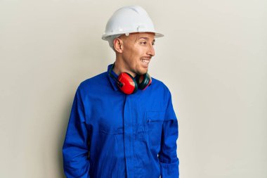 Sakallı, inşaatçı tulumu giyen ve yüzünde gülümsemeyle yan yana duran kel bir adam, doğal bir ifade. Kendinden emin gülen. 