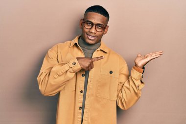 Genç siyahi adam gündelik kıyafetler ve gözlük takıyor kameraya hayret ediyor ve gülümsüyor.. 