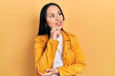 Burnunda piercing olan, elleri çenesinde sarı deri ceket giyen soru soran, düşünceli bir İspanyol kadın. Gülümseyen ve düşünceli bir yüz. şüphe kavramı. 