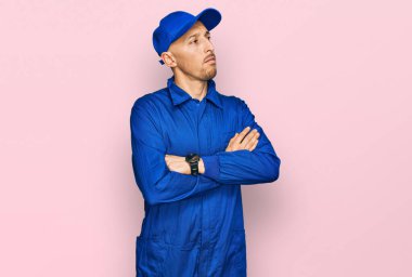 Kel, sakallı, işçi tulumu giyen, kolları çapraz ve kendinden emin bir şekilde yana bakan bir adam. 