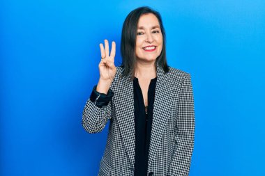 Orta yaşlı İspanyol kadın iş kıyafetleri giyip üç numaralı parmaklarıyla işaret ederken kendinden emin ve mutlu bir şekilde gülümsüyor.. 