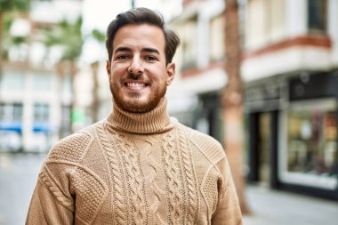 Genç beyaz adam gülümseyerek şehirde mutlu mesut duruyor..