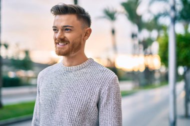 Genç beyaz adam gülümseyerek şehirde mutlu mesut duruyor..