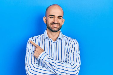 Zarif kıyafetler giymiş genç kel bir adam. Gülümsüyor. Elleri ve parmakları yana doğru uzanıyor. 