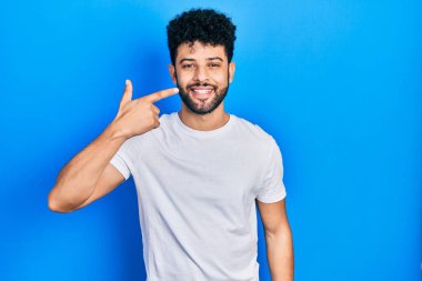 Sakallı, günlük beyaz tişörtlü, el ve yüz parmaklarını ve burnunu gösteren, neşeli gülümseyen genç bir Arap. güzellik konsepti 