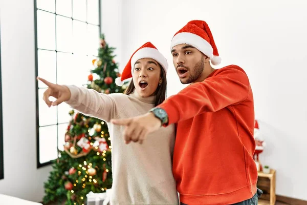 Jovem Casal Hispânico Junto Árvore Natal Apontando Com Dedo Surpreso — Fotografia de Stock