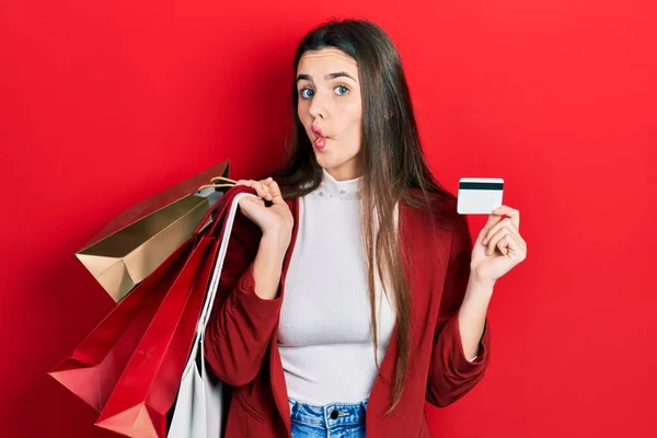 Young brunette teenager holding shopping bags and credit card making fish face with mouth and squinting eyes, crazy and comical.