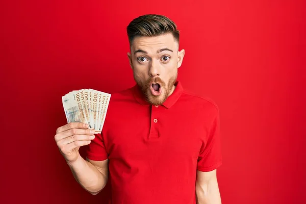 Young Redhead Man Holding 100 Danish Krone Banknotes Scared Amazed —  Fotos de Stock