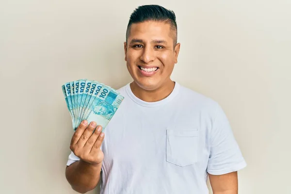 Young Latin Man Holding 100 Brazilian Real Banknotes Looking Positive — 图库照片