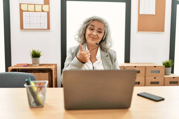 Middle age businesswoman sitting on desk working using laptop at office doing happy thumbs up gesture with hand. approving expression looking at the camera showing success.