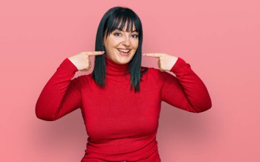 Günlük kıyafetler giyen, neşeli gülümseyen, parmaklarıyla ve ağzıyla işaret eden genç İspanyol kadın. diş sağlığı kavramı. 