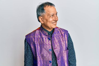 Orta yaşlı Hintli bir adam geleneksel Hint kıyafetleri giyiyor. Tarafsız, rahat bir görünüm takınıyor. Kendine güvenen bir gülümsemesi var.. 