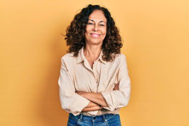 Orta yaşlı İspanyol kadın gündelik kıyafetler giyip kollarını kavuşturup kameraya bakarken gülümsüyor. pozitif kişi. 