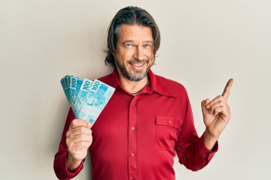 Orta yaşlı yakışıklı adam elinde 100 Brezilyalı gerçek banknot tutarken mutlu bir şekilde elini ve parmağını kenarda gezdiriyor. 