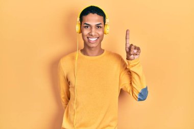 Genç Afro-Amerikalı adam müzik dinlerken kulaklık takıyor ve bir numaralı parmağını gösterip işaret ederken kendinden emin ve mutlu gülümsüyor.. 