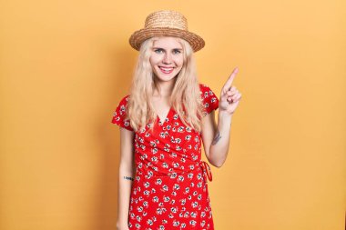 Sarışın, güzel, beyaz bir kadın. Yaz şapkası takıyor. Bir numaralı parmağını gösterip gülümsüyor.. 