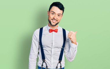 Hipster sakallı İspanyol adam papyonlu ve pantolon askısı takan mutlu yüzlü gülümseyen ve başparmağıyla yan tarafı gösteren.. 