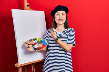 Orta yaşlı İspanyol kadın paletle resim çiziyor, resim sehpası, neşeli duruyor, yüzünde gülümseme, el ve parmaklarıyla işaret ediyor, mutlu ve doğal bir ifadeyle. 