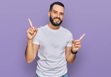 Sakallı, günlük beyaz tişörtlü, kendine güvenen, parmakları farklı yönleri gösteren genç bir adam. reklam için kopyalama alanı 