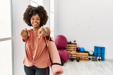 Afro saçlı, pilates odasında yoga minderi tutan Afro-Amerikalı bir kadın seni ve kamerayı parmaklarıyla göstererek pozitif ve neşeli bir şekilde gülümsüyor. 