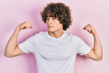 Sıradan beyaz tişört giyen yakışıklı genç adam gururla gülümseyen kol kaslarını gösteriyor. fitness konsepti. 