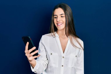 Genç esmer kız akıllı telefon kullanıyor. Masada otururken mutlu ve pozitif görünüyor. Kendine güvenen bir gülümsemeyle dişlerini gösteriyor. 