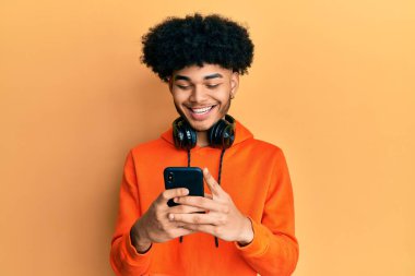 Afro saçlı, mutlu ve havalı bir gülümsemeyle gülümseyen akıllı telefon kullanan Afro-Amerikan genç adam. diş gösterme. 