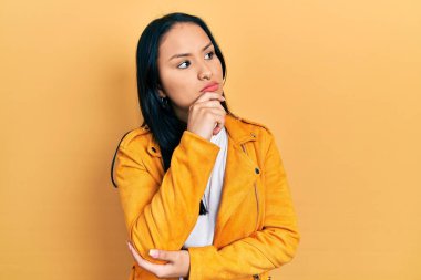 Burnunda piercing olan, elleri çenesinde sarı deri ceket giyen soru soran, düşünceli bir İspanyol kadın. Düşünceli bir yüzle gülümsüyor. şüphe kavramı. 