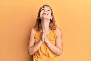 Kolsuz tişörtlü, sıradan giyinen genç beyaz bir kadın elleri bağlı yalvarıyor ve dua ediyor. Yüzünde umut ifadesi var. Çok duygusal ve endişeli. Yalvarıyor. 