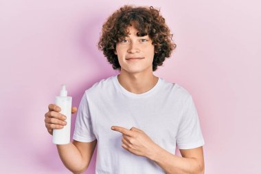 Kozmetik nemlendirici krem tutan yakışıklı genç adam gülümseyerek mutlu bir şekilde eli ve parmağı işaret ediyor. 