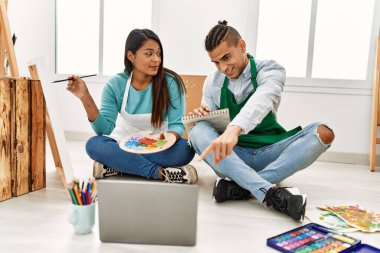 Genç Latin ressam çift, resim stüdyosunun zemininde dizüstü bilgisayar kullanarak çevrimiçi boya dersi alıyorlar..