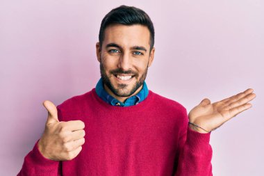 Latin asıllı genç adam, günlük kıyafetler giymiş, avuç içini gösteriyor ve baş parmaklarıyla iyi bir jest yapıyor, mutlu ve neşeli gülümsüyor. 