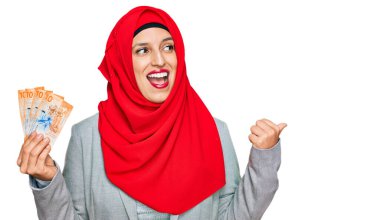 İslami başörtüsü takan güzel İspanyol kadın elinde 10 İsviçre frangı banknotuyla baş parmağını yan tarafa uzatarak mutlu bir şekilde gülümsüyor. 