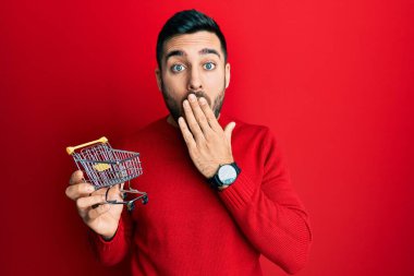 Küçük bir süpermarket alışveriş arabası tutan genç İspanyol adam ağzını eliyle kapatıyor, şokta ve hata yapmaktan korkuyor. şaşırmış ifade 