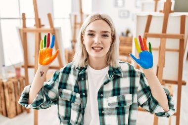 Resim stüdyosunda renkli boyalı eller göstererek mutlu gülümseyen genç sanatçı kız..