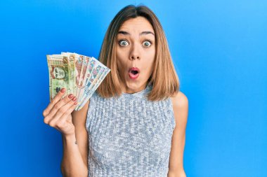 Genç, beyaz, sarışın bir kadın elinde Peru banknotları tutuyor. Korkmuş ve şaşırmış bir şekilde ağzı açık, inançsız bir yüz. 