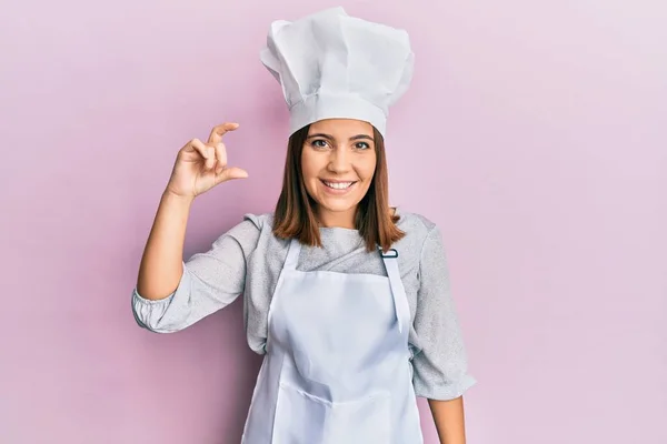 年轻漂亮的女人穿着职业厨师制服 戴着帽子 微笑着 自信地做手势 手拿着小尺寸的标志 手拿着手指 手拿着相机 计量概念 — 图库照片