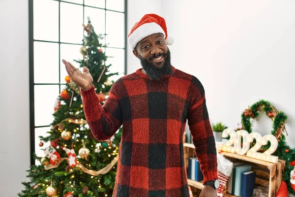 アフリカ系アメリカ人男性身に着けているサンタクラスの帽子立っているクリスマスツリー笑顔陽気なプレゼンテーションとポインティングで手の手のひらカメラを見て — ストック写真