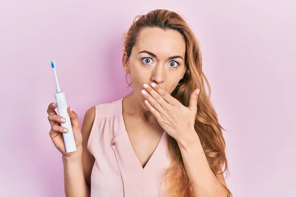 Young caucasian woman holding electric toothbrush covering mouth with hand, shocked and afraid for mistake. surprised expression