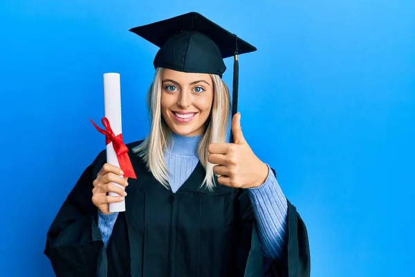 卒業キャップと式のローブを身に着けている美しいブロンドの女性が学位を保持笑顔幸せと正 親指アップ行う優れた承認サイン — ストック写真